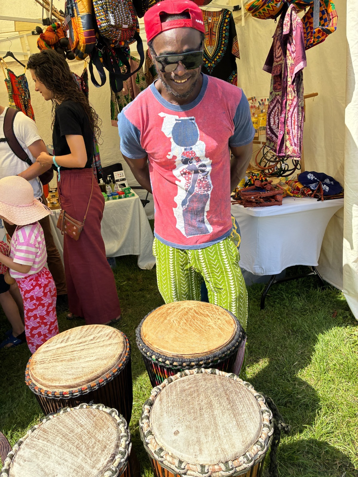 Vendor in red cap and sunglasses behind four djembe drums inside the tent
